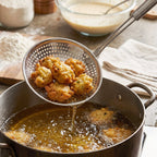 A stainless steel perforated skimmer spoon with a long handle, lifting fried fritters from a pot of hot oil.