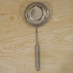 A stainless steel perforated skimmer spoon with a long handle resting on a light wooden surface.