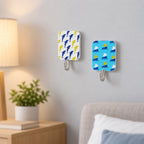 Dolphin and sailboat patterned hooks affixed to a light gray wall above a bedside table with a lamp and plant.