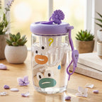 A clear glass tea sipper cup with a purple lid and cartoon duck face design, filled with water, sits on a wooden table with