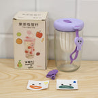 A glass tea sipper cup with a purple lid, a box, and two cartoon face stickers are displayed on a wooden surface.