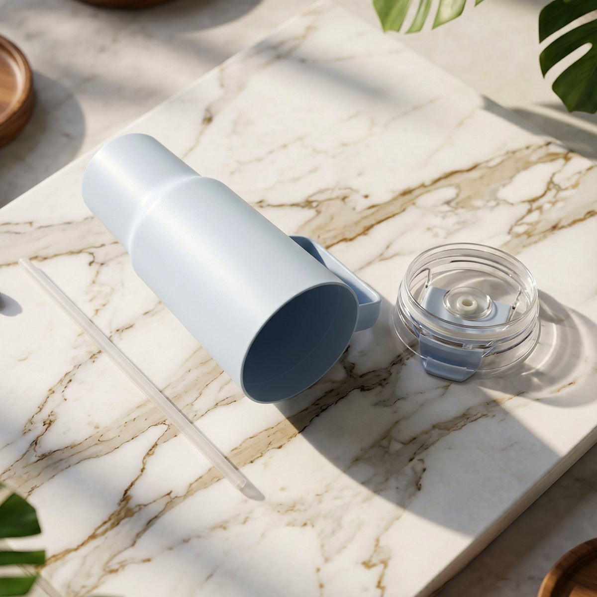 A light blue handled tumbler lies on its side on a marble surface with its clear lid and straw beside it.