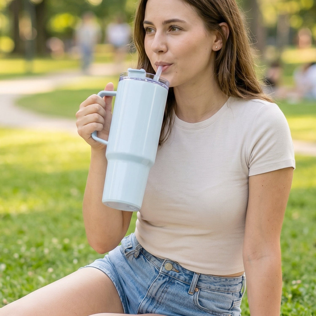 A woman sits on grass, sipping from a large light blue handled tumbler with a straw.