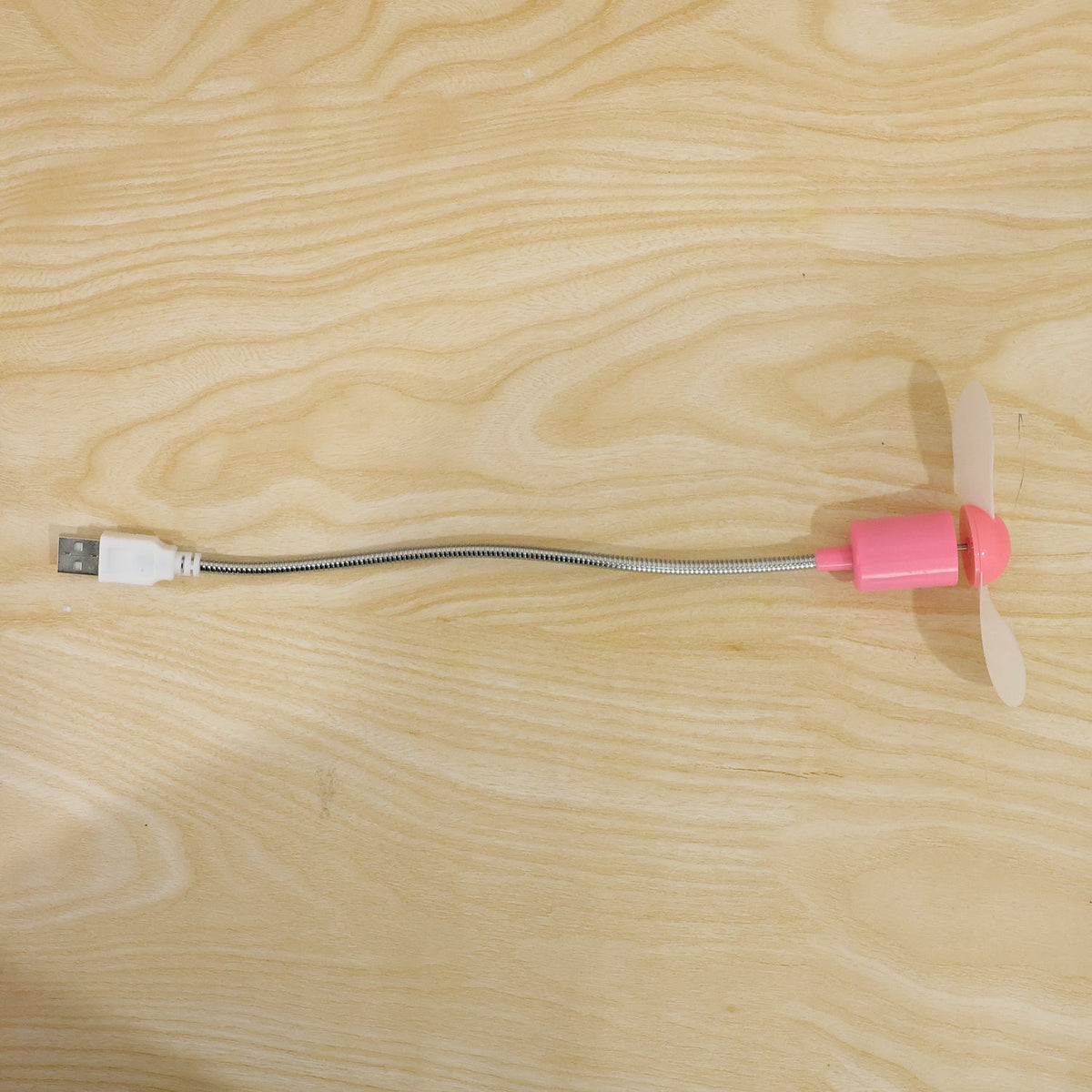 Pink USB mini fan with white blades and a flexible silver gooseneck, laid flat on a light wooden surface.