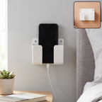 A white wall-mounted phone holder with a black smartphone charging in a bedroom setting.