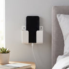 A white wall-mounted phone holder with a black smartphone charging next to a bed and nightstand.