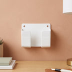 A white wall-mounted phone holder with two compartments on a peach-colored wall above a wooden desk.