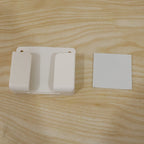 A white wall-mounted phone holder and a small white square adhesive pad on a light wooden surface.