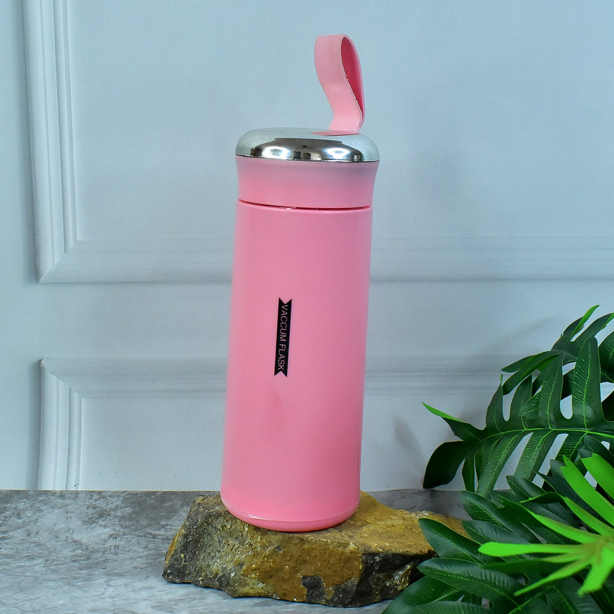 A pink vacuum flask with a silver lid and strap sits on a rock with green leaves and a white wall in the background.