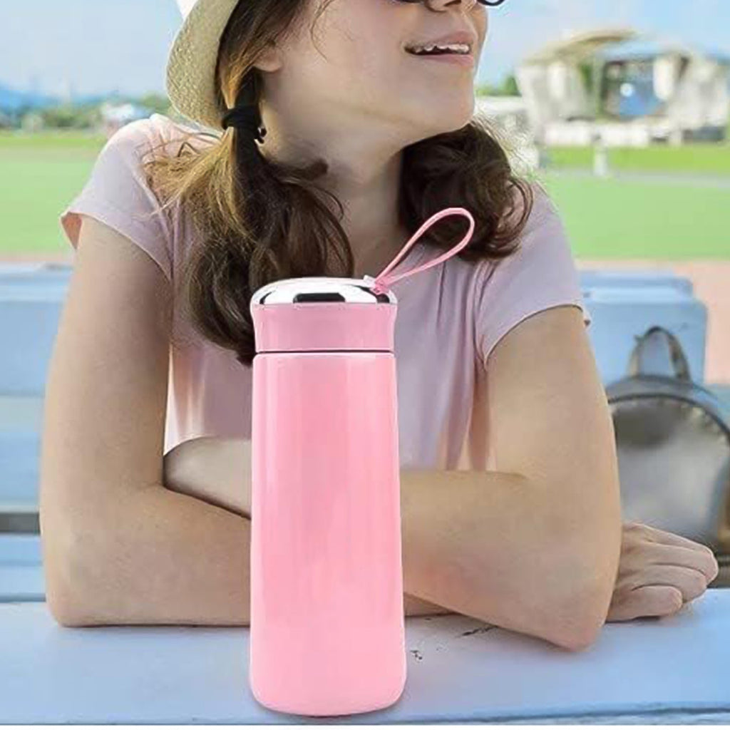 A pink vacuum flask with a silver lid and strap sits on a table in front of a person wearing a straw hat.