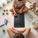 Two children sit on a play mat, one writing "ABC 123" on a black LCD tablet, surrounded by toys and books.