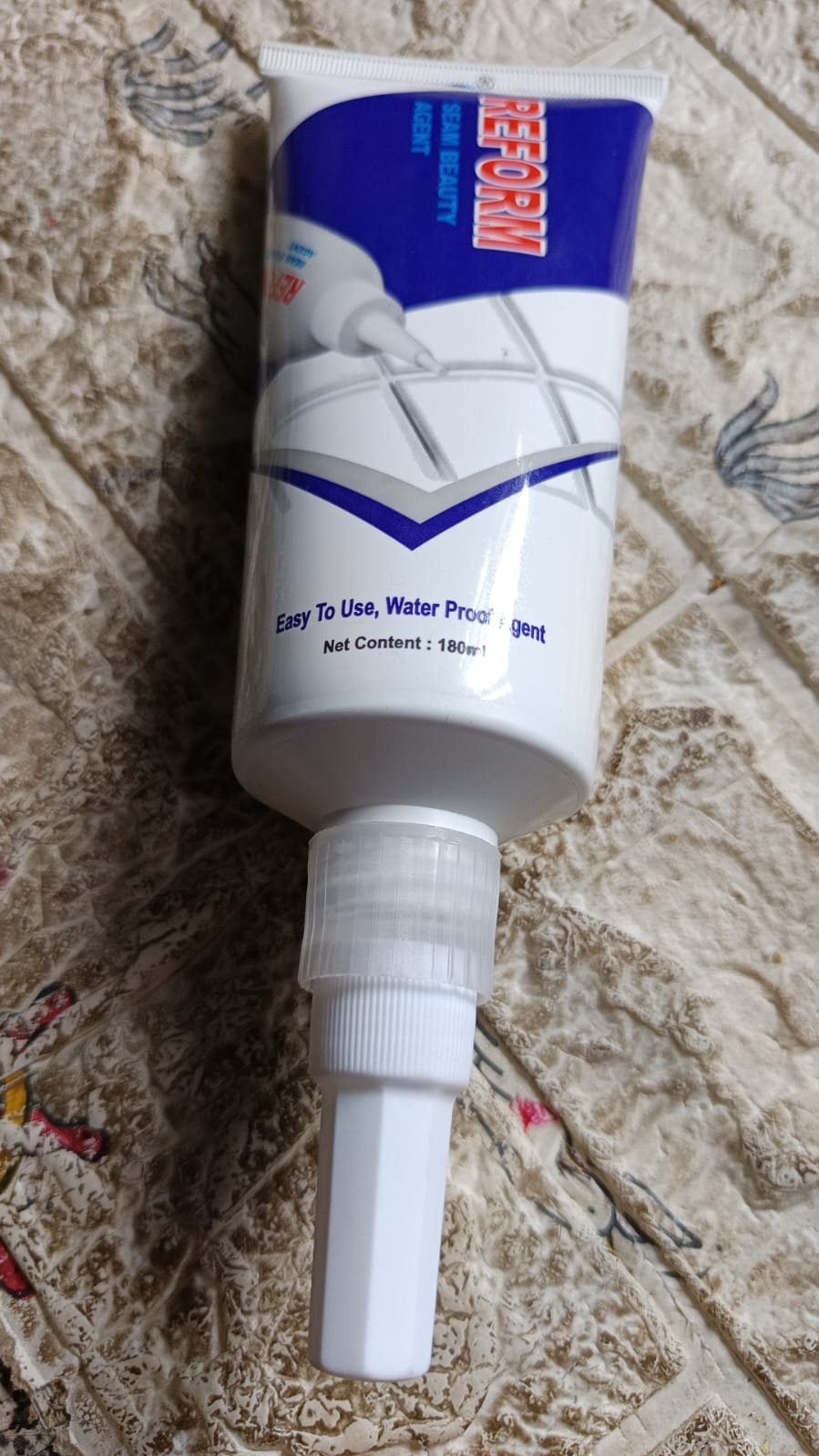 A white and blue tube of 'Seam Beauty Agent' sealant with a long nozzle, resting on a textured brown surface.