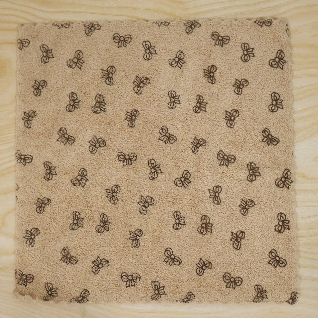 A beige microfiber towel with a dark bow pattern and scalloped edges lies flat on a light wood surface.