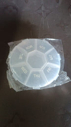 A clear 7-day pill organizer, still in its plastic packaging, on a dark surface.