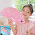 A young girl in a pink dress smiles while holding an open pink folding fan decorated with white flowers and text.