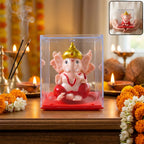 A pink Ganesha idol with a gold crown sits in a clear plastic box on a red base, surrounded by lit diyas and flowers.