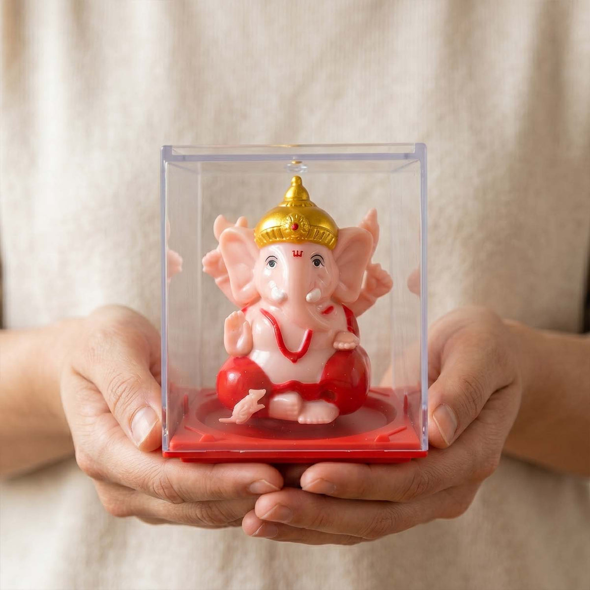 Two hands gently hold a clear plastic box containing a pink Ganesha idol with a gold crown on a red base.