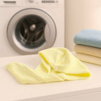 A yellow microfiber hair wrap towel with a button is placed on a white surface, with a washing machine and folded towels in