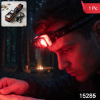 A man wearing an orange and black headlamp with a red light, reading a map in a dark forest with a campfire in the – view 2