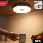 A hand reaches for a mug under a bright, round LED light mounted beneath a wooden cabinet, illuminating a fruit bowl