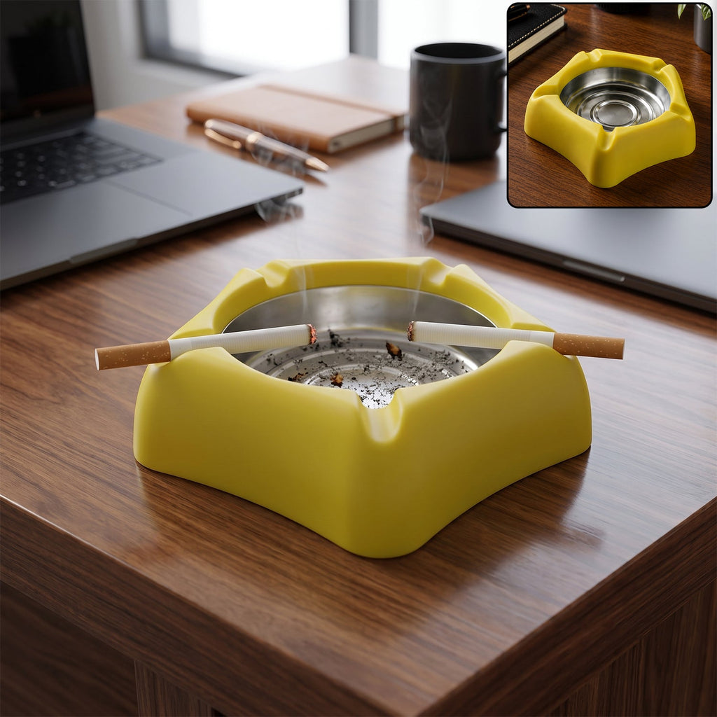 A yellow silicone ashtray with a stainless steel insert, holding two lit cigarettes, sits on a wooden desk with laptops.