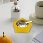 A yellow silicone ashtray with a stainless steel insert, holding one lit cigarette, sits on a white table next to a cup and