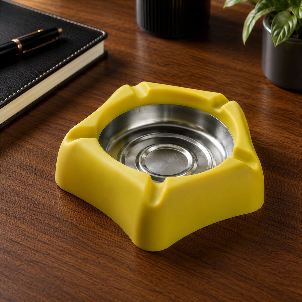 A yellow silicone ashtray with a stainless steel insert is positioned on a dark wooden desk next to a black notebook.