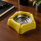 A yellow silicone ashtray with a stainless steel insert is positioned on a dark wooden desk next to a black notebook.