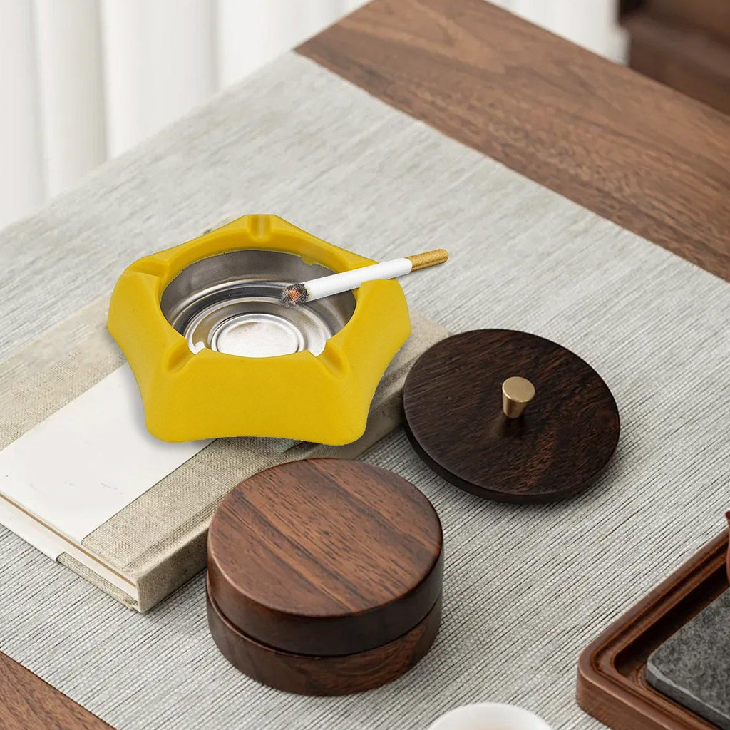 A yellow silicone ashtray with a lit cigarette sits on a light grey placemat, next to two dark wooden containers.